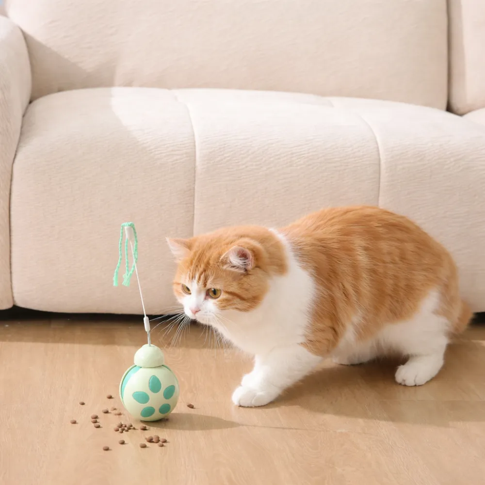 An orange and white ginger cat pouncing toward a green treat dispenser toy with kibble scattered on the floor.
