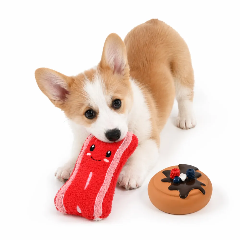 corgi-puppy-plush-bacon-dog-toy.jpg A cute Corgi puppy lying down and biting a red smiling plush bacon dog toy next to a latex donut toy.