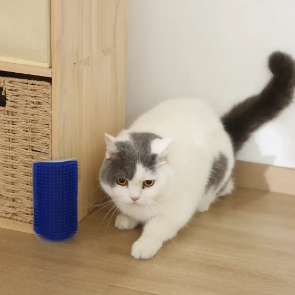 A grey and white cat with bright eyes crouches on a wooden floor, pawing and rubbing its chin against a detached blue Cat Self-Groomer brush. The cat's long, dark tail is curved gracefully behind it. A woven basket and cabinet are in the background.