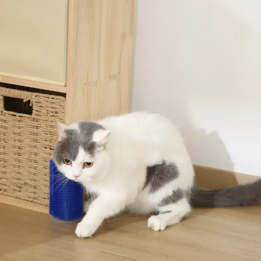 A curious grey and white cat approaches and uses a blue Cat Self-Groomer attached to the corner of a light wood furniture leg. The cat rubs its cheek against the soft silicone bristles. A basket and cabinet are visible in the background.