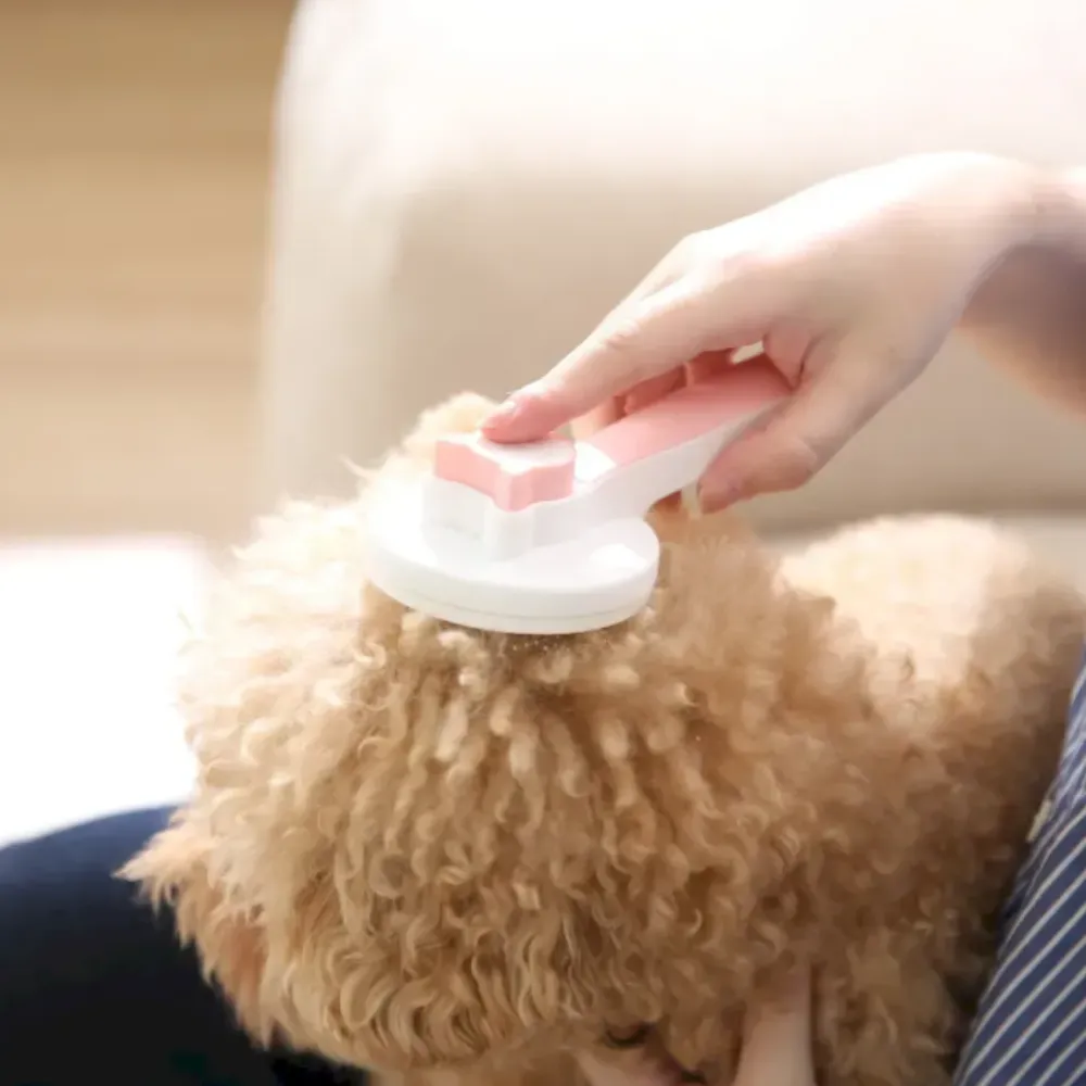 Close-up of a pink pet grooming brush being used on tan dog fur to demonstrate gentle de-shedding.