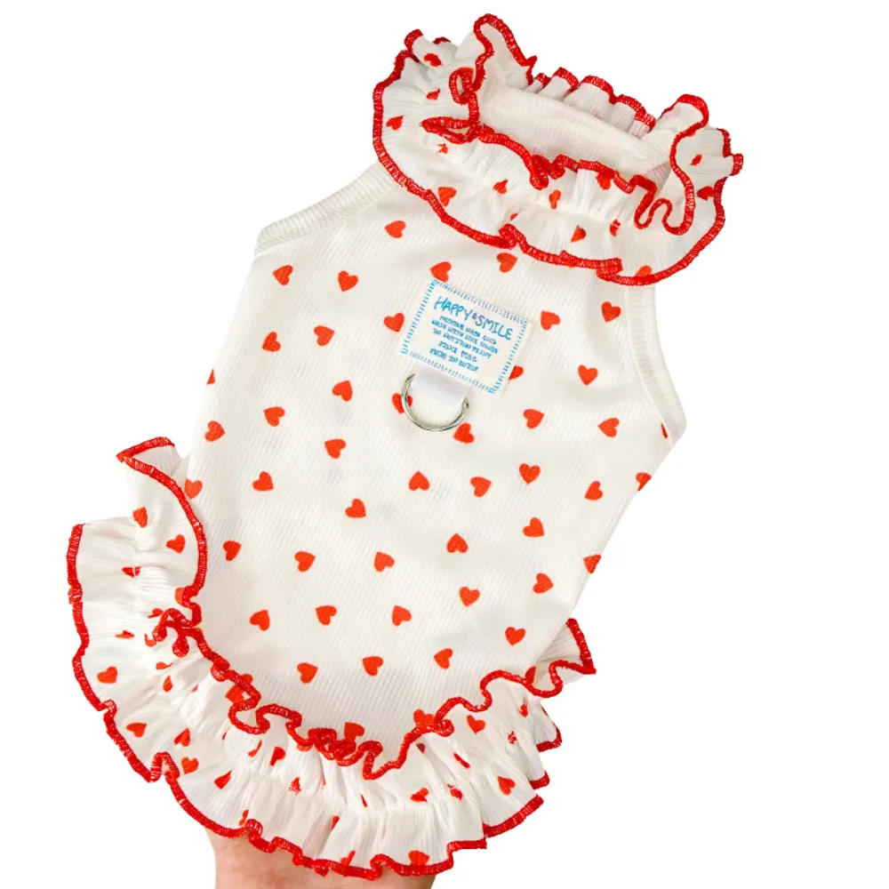 Close-up of a white dog dress with red heart prints, featuring a ruffled collar and a built-in metal D-ring for leash attachment.